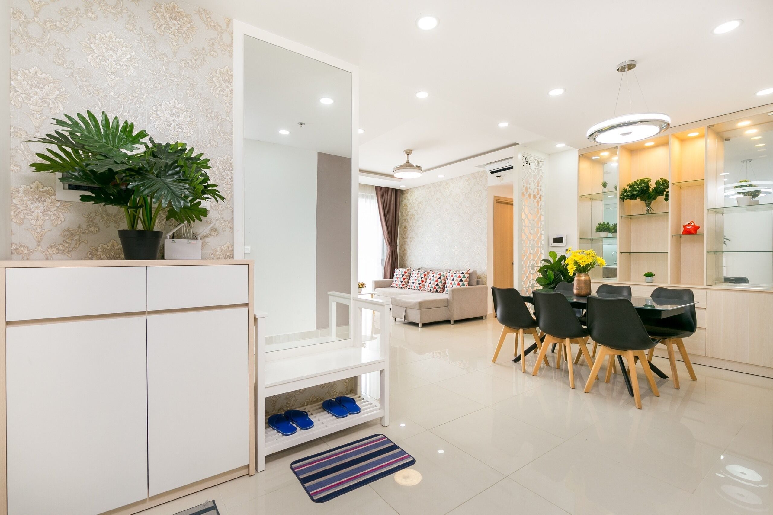 A modern living room with a sofa, dining table, and chairs. There is a potted plant and a shoe rack.