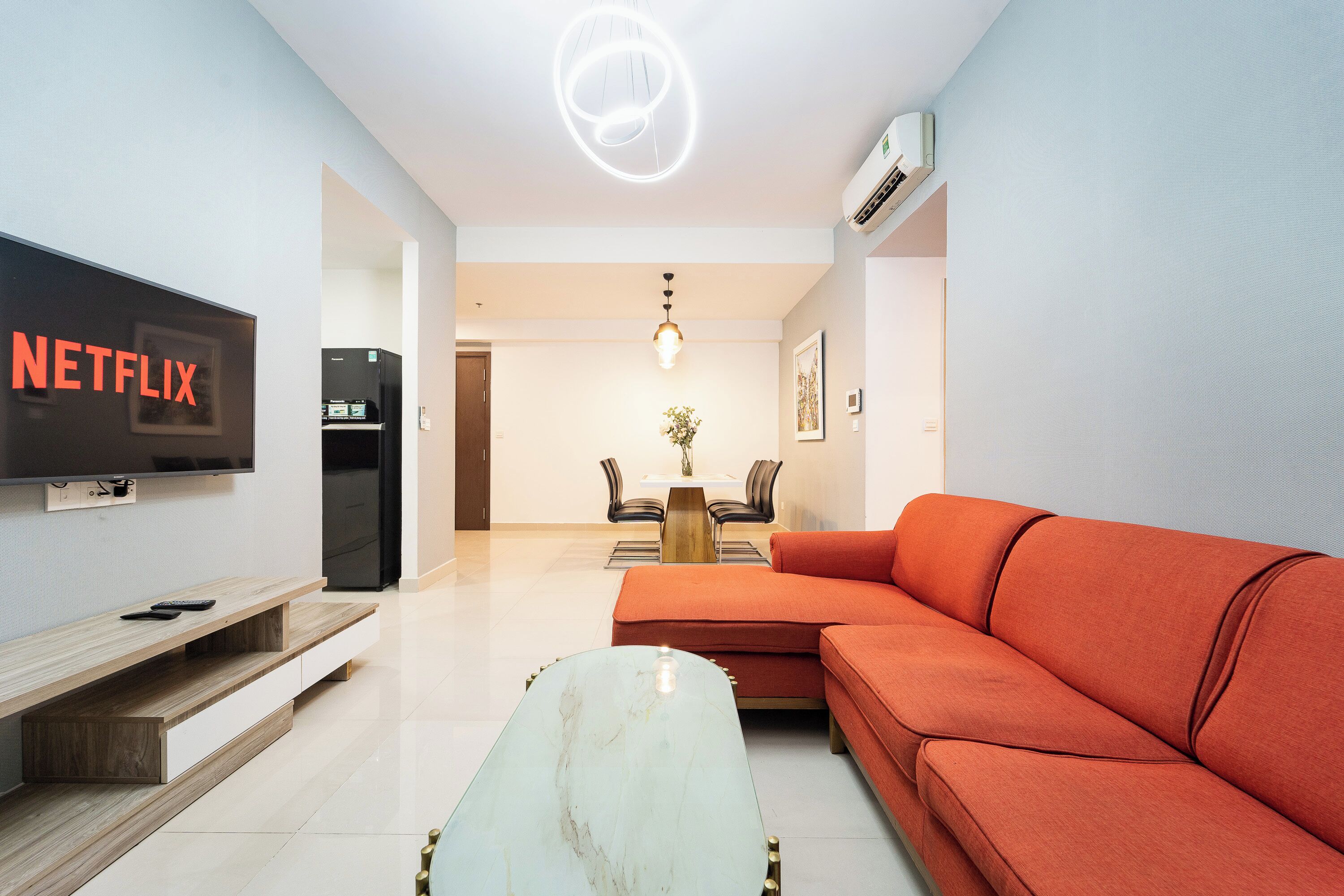 A modern living room with a flat-screen TV, a sofa, a coffee table, and a dining area in the background.