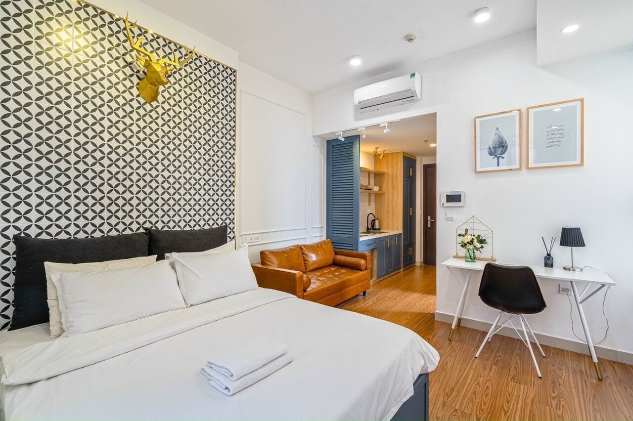A modern hotel room with a bed, a brown leather sofa, a desk with a chair, and a wall-mounted air conditioner.