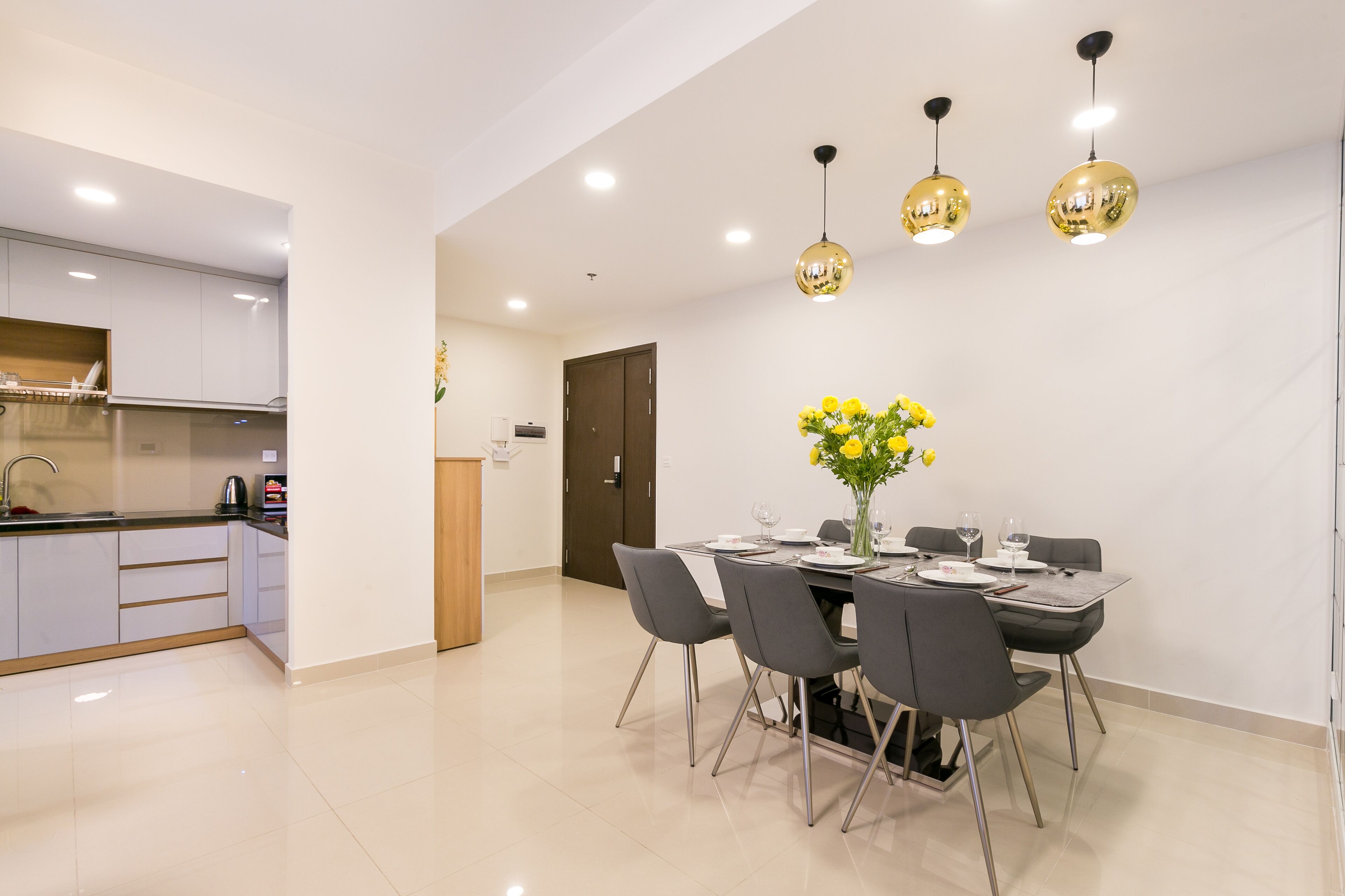 A modern kitchen with white cabinets, a black countertop, and a built-in oven. There is a dining area with a table set for four, featuring a vase with yellow flowers.