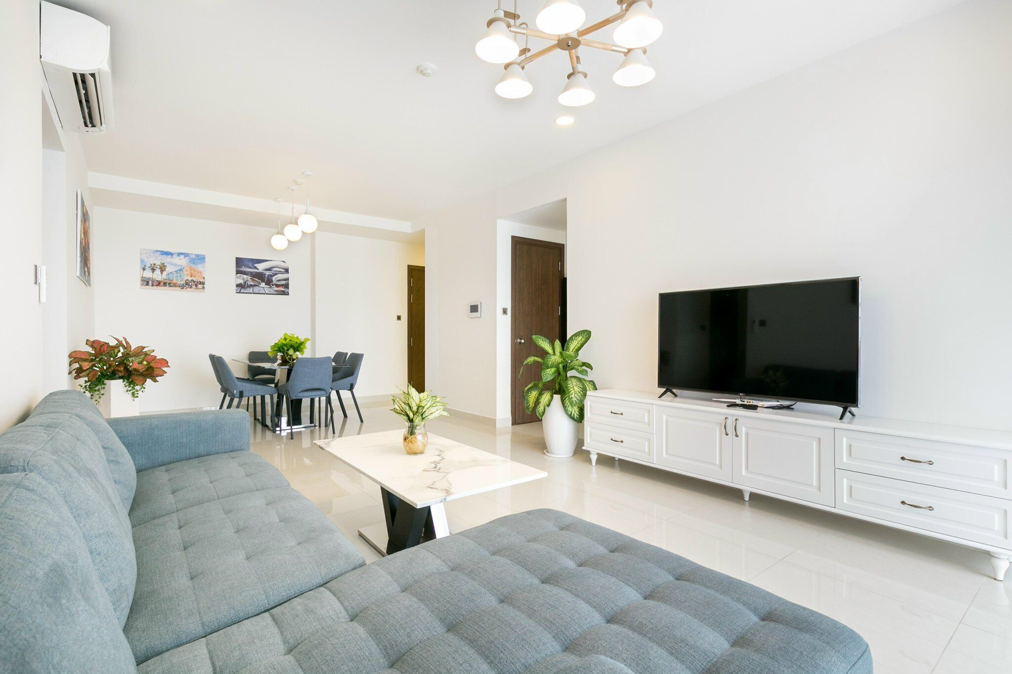 A modern living room with a grey sofa, a dining area with chairs and a table, a TV on a white cabinet, and a ceiling light fixture.