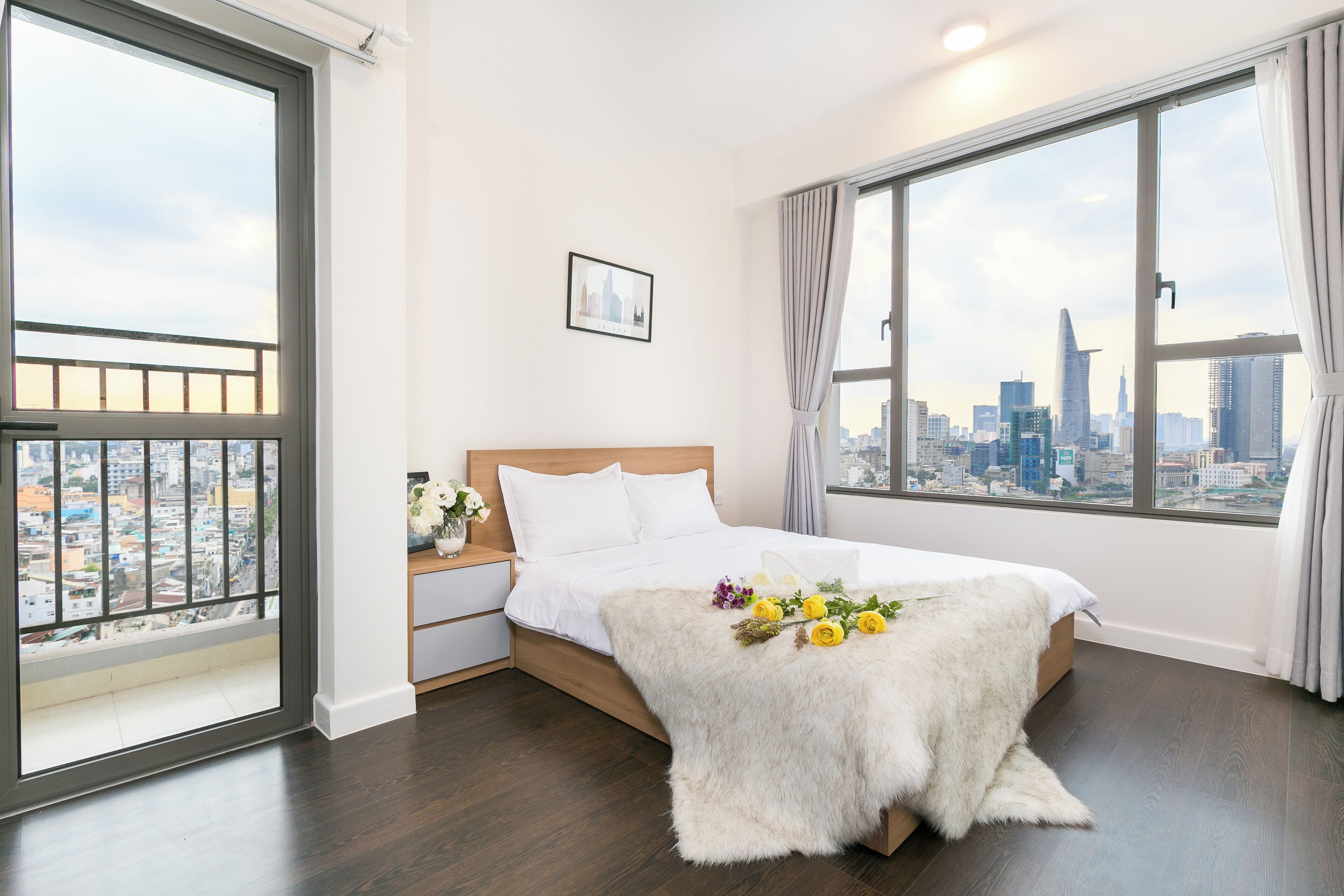 A bedroom with a city view, a bed with a white blanket, a nightstand, and a window with curtains.