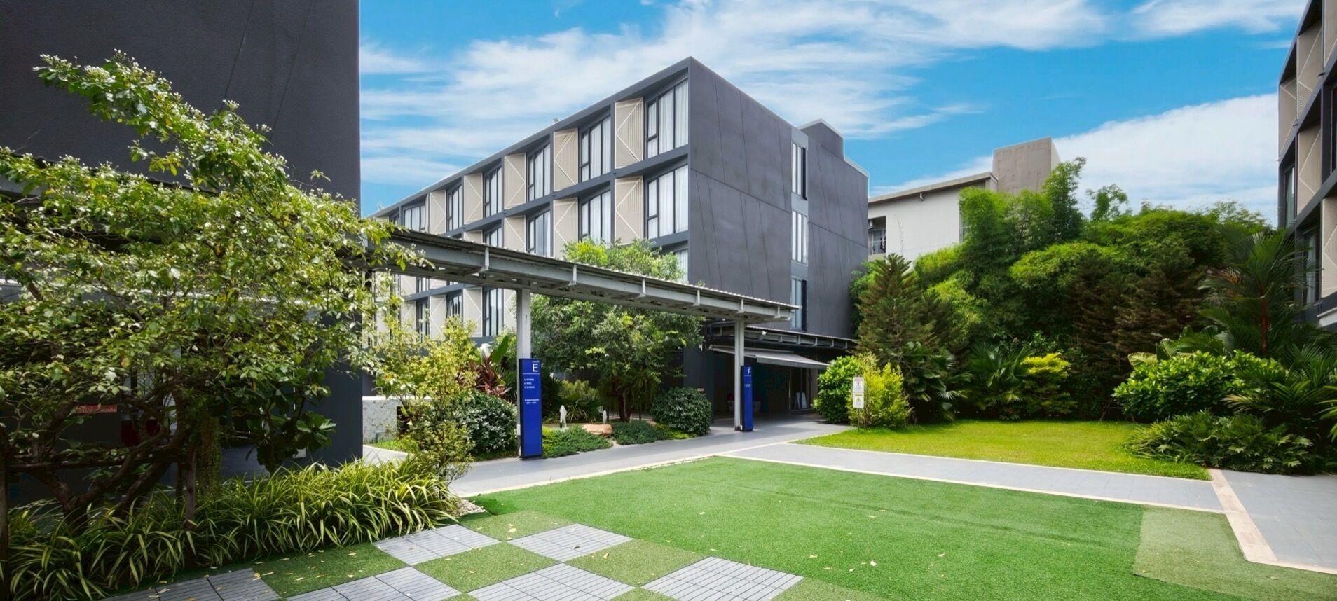 Un edificio moderno con un paseo techado, césped verde y diversas plantas.