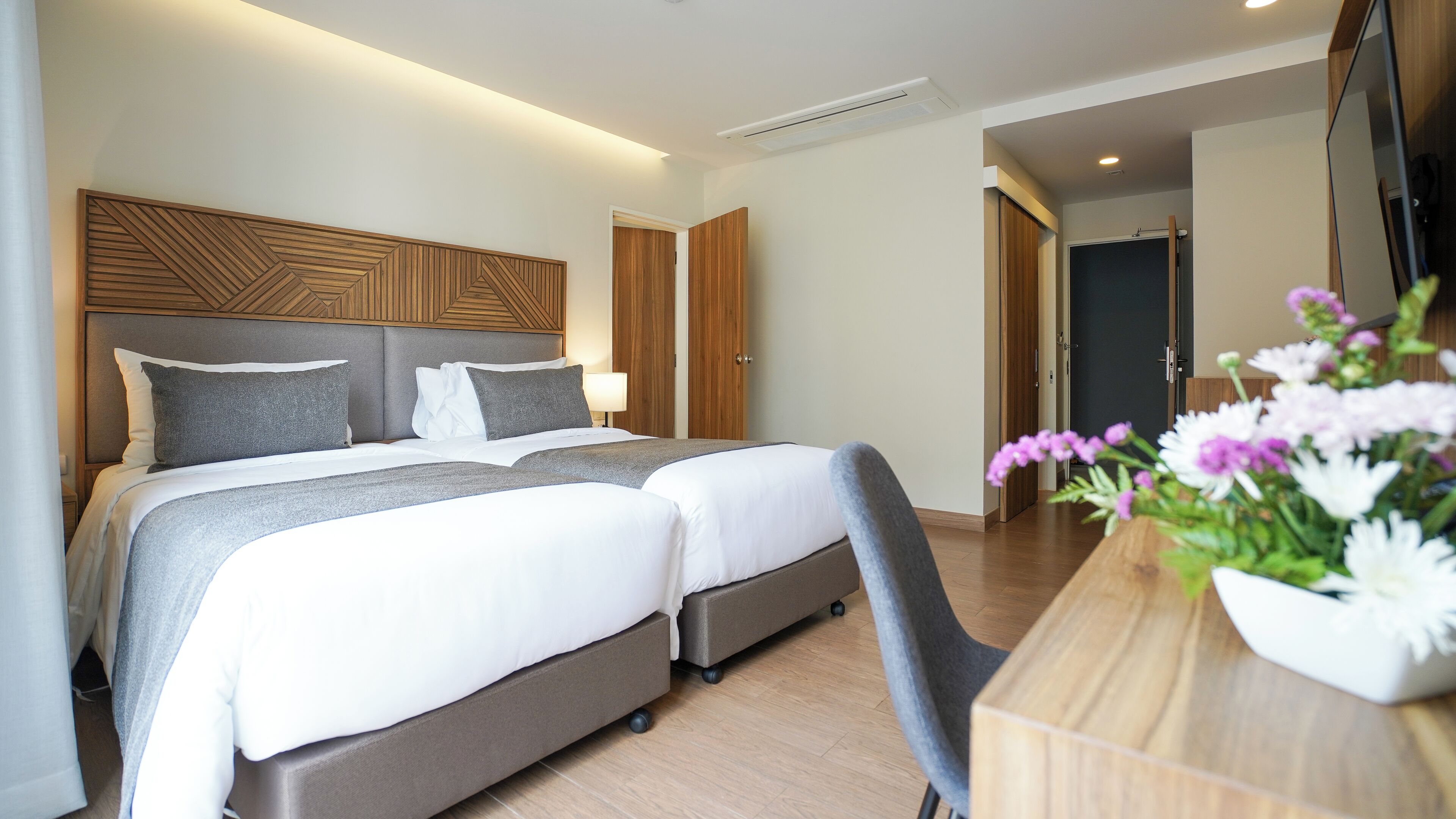 Een moderne hotelkamer met een groot bed, een houten hoofdbord, een bureau met stoel en een bloevas.