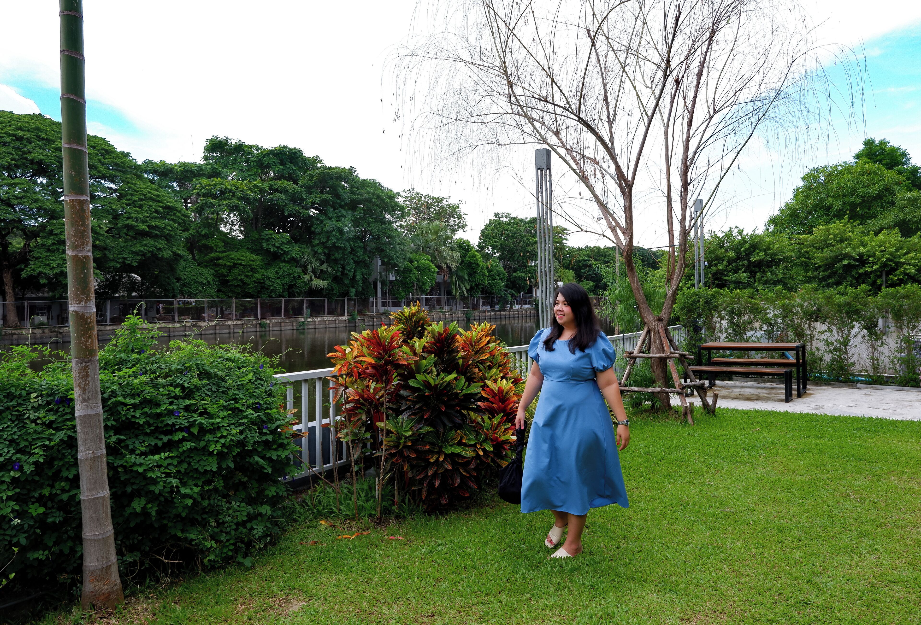 Una mujer con un vestido azul parada en un parque, con un banco y un árbol de fondo.