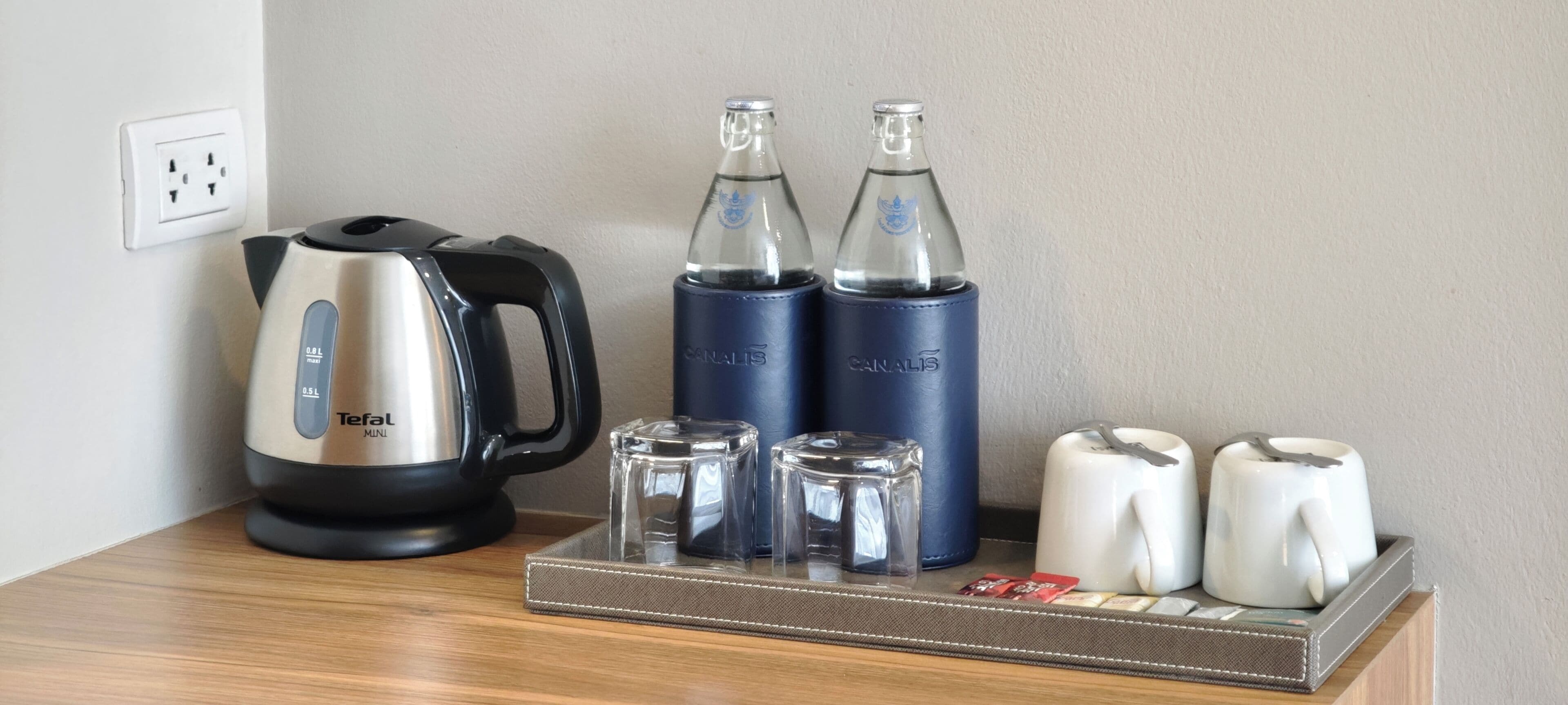Una kitchenette con un hervidor Tefal Mini, dos botellas de agua azules, una jarra de vidrio y un juego de tazas blancas sobre una bandeja.