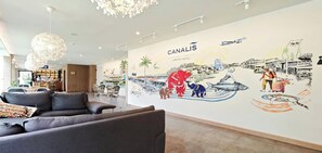 A hotel lobby with a mural depicting Bangkok's landmarks and activities, a reception area with a sofa and chairs, and a chandelier.