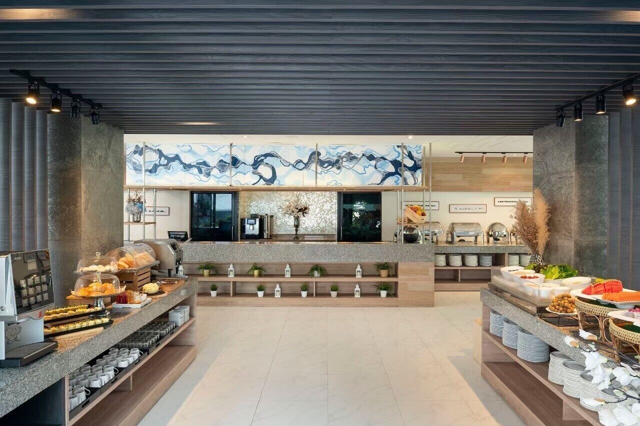 A modern hotel lobby with a central reception desk, a decorative mural, and a buffet area with various food items.