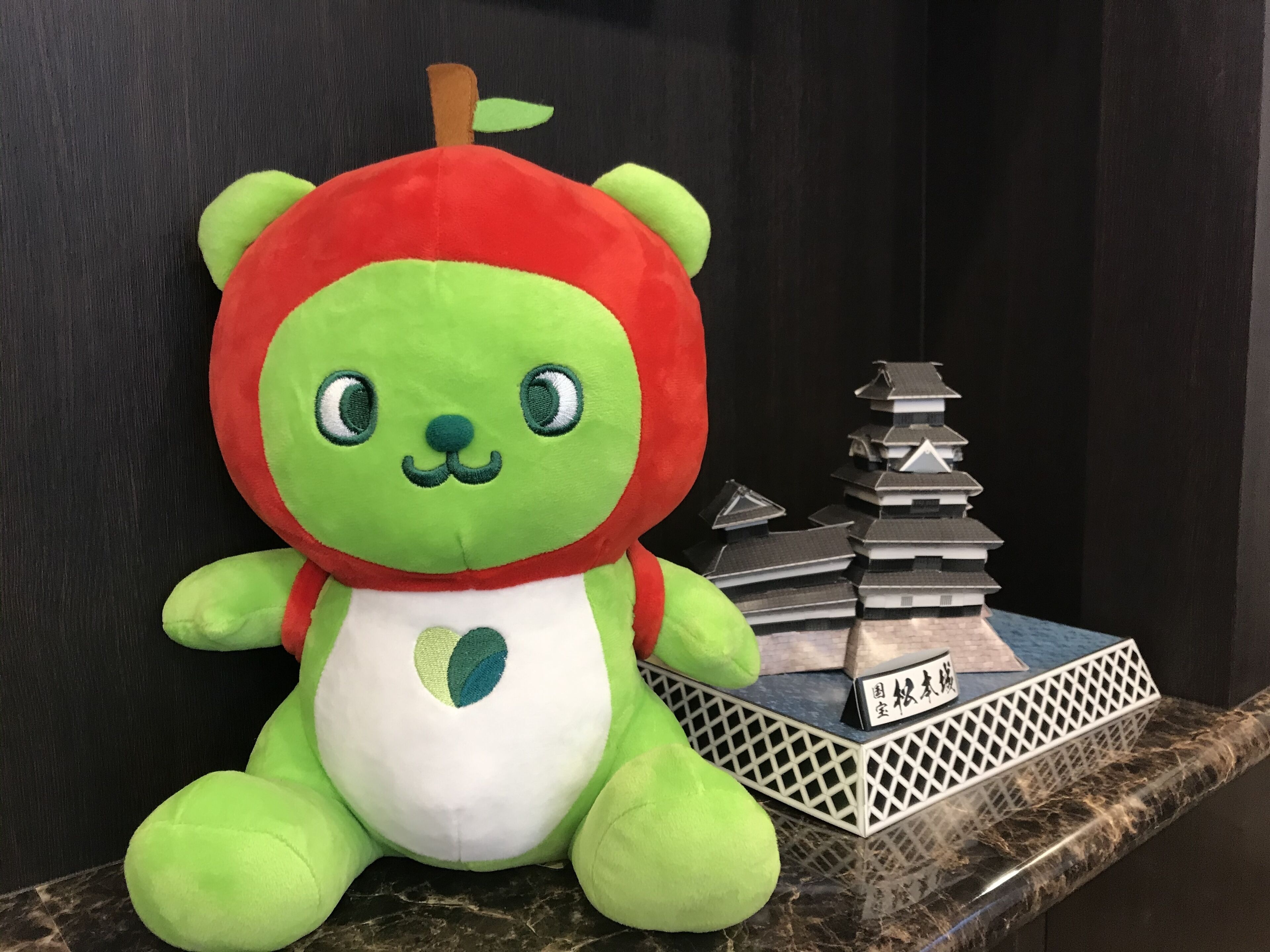 A plush toy with a red and green head, sitting on a marble surface next to a stack of books and a small model of a pagoda.
