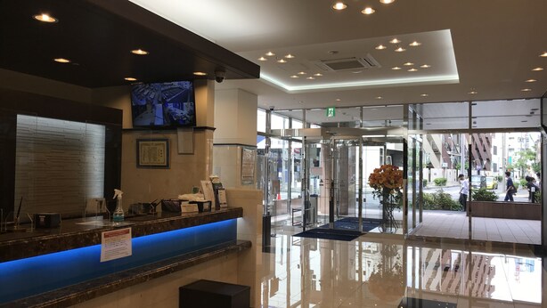 A hotel reception area with a blue-lit counter, a mounted television, and a glass entrance leading outside. - Toyoko Inn Matsumoto-eki Higashi-guchi (Matsumoto)
