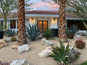 Une maison avec un toit en tuiles, entourée de différentes espèces de cactus et de plantes d’agave, et une allée gravillonnée.