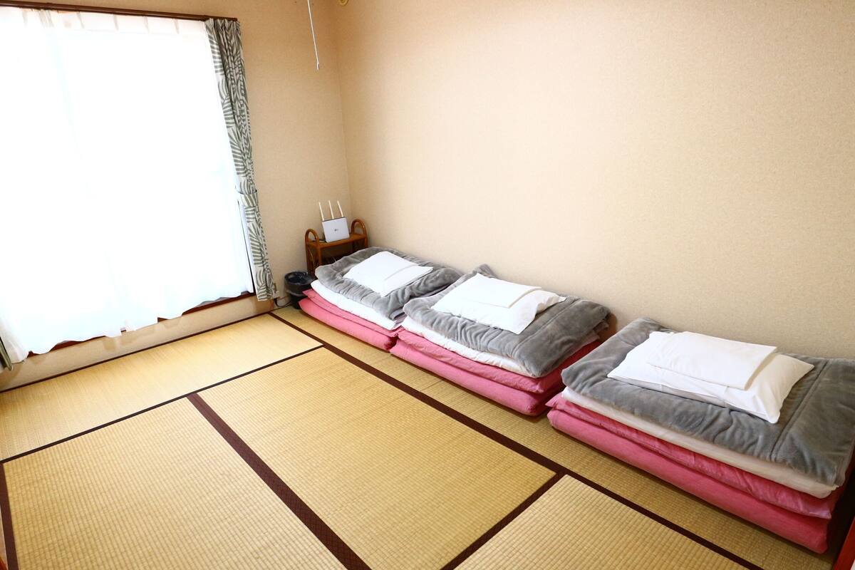 Una habitación con suelo de esteras de tatami, dos camas con almohadas, un ordenador portátil sobre una mesita auxiliar y una ventana con cortinas.