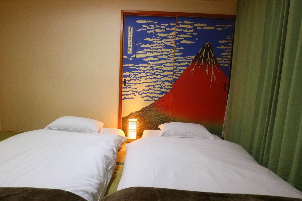 Habitación de hotel con dos camas, una pared con un mural de montañas y nubes, y una cortina verde.