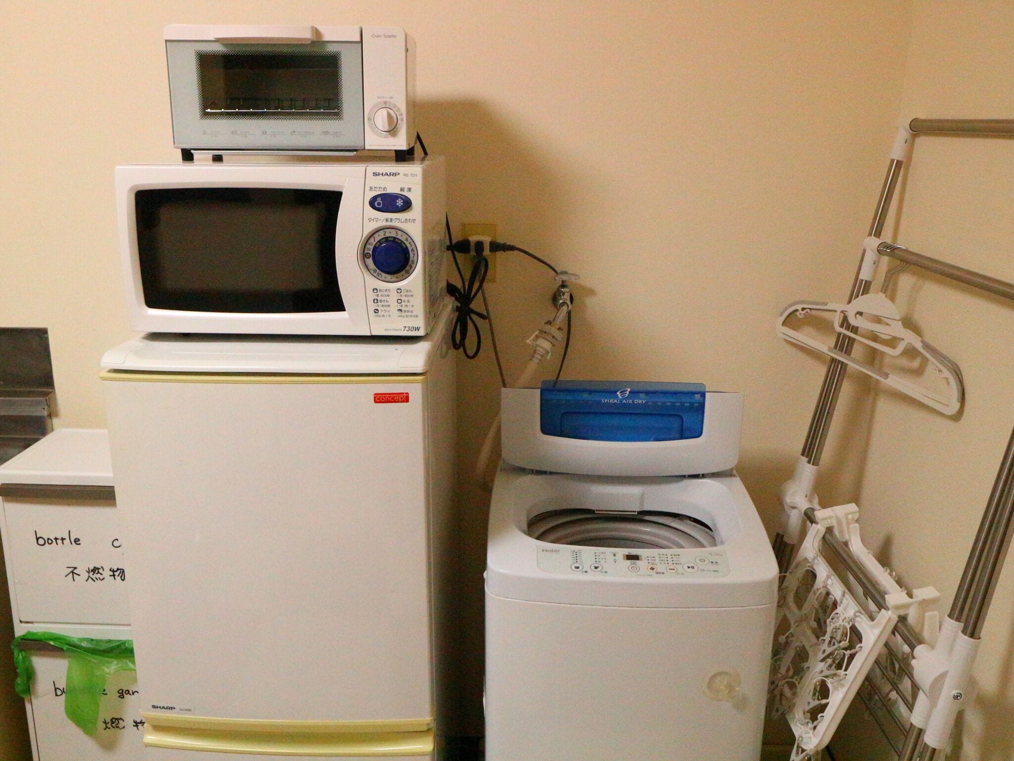 Una habitación compacta con microondas sobre refrigerador, lavadora y un estante metálico con una toalla blanca.