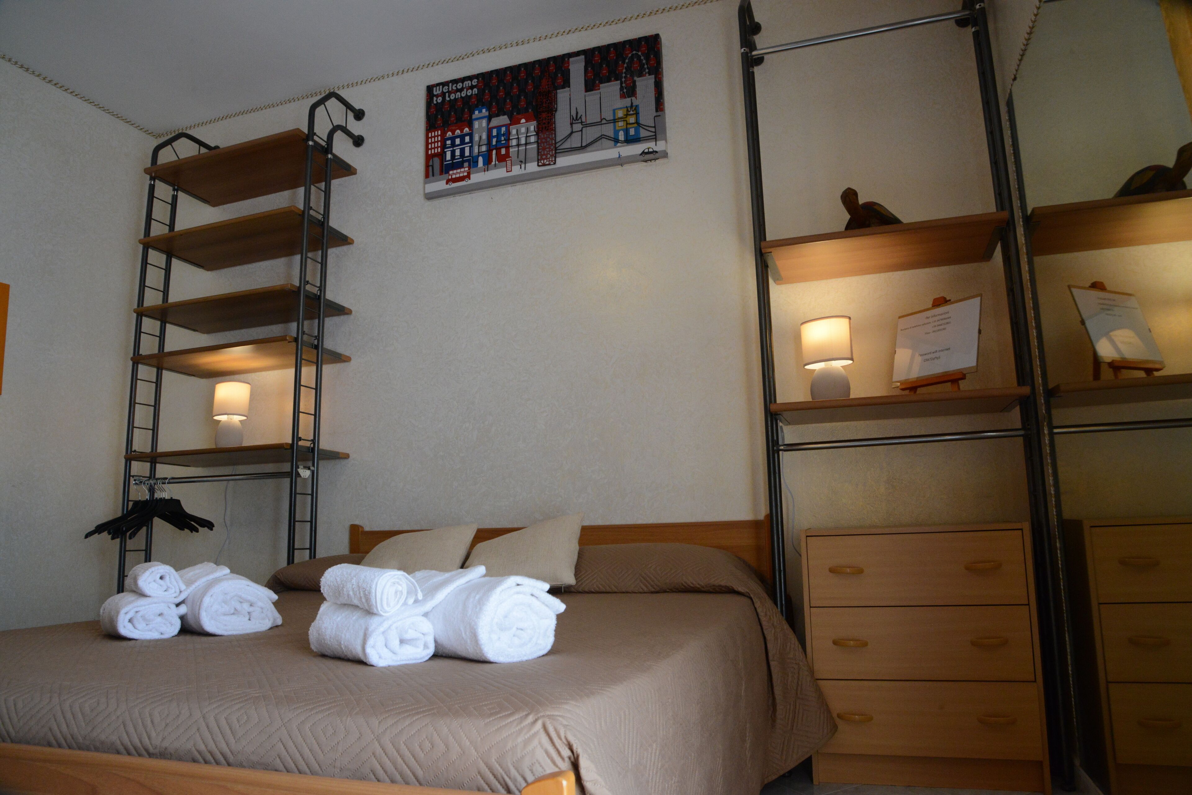 A neatly made bed with rolled towels, a ladder, and a wall-mounted shelf with a lamp and papers.