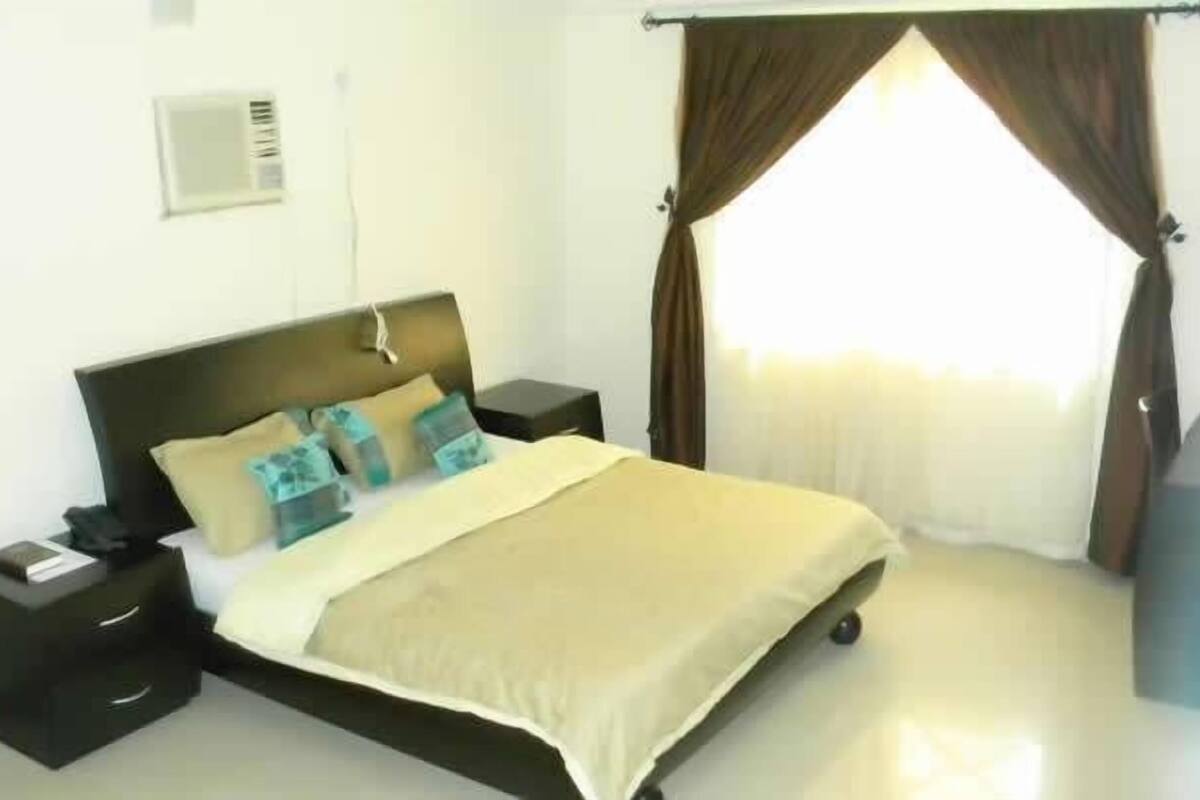 A bedroom with a bed, nightstands, a bedside table, a window with curtains, and a wall-mounted thermostat.