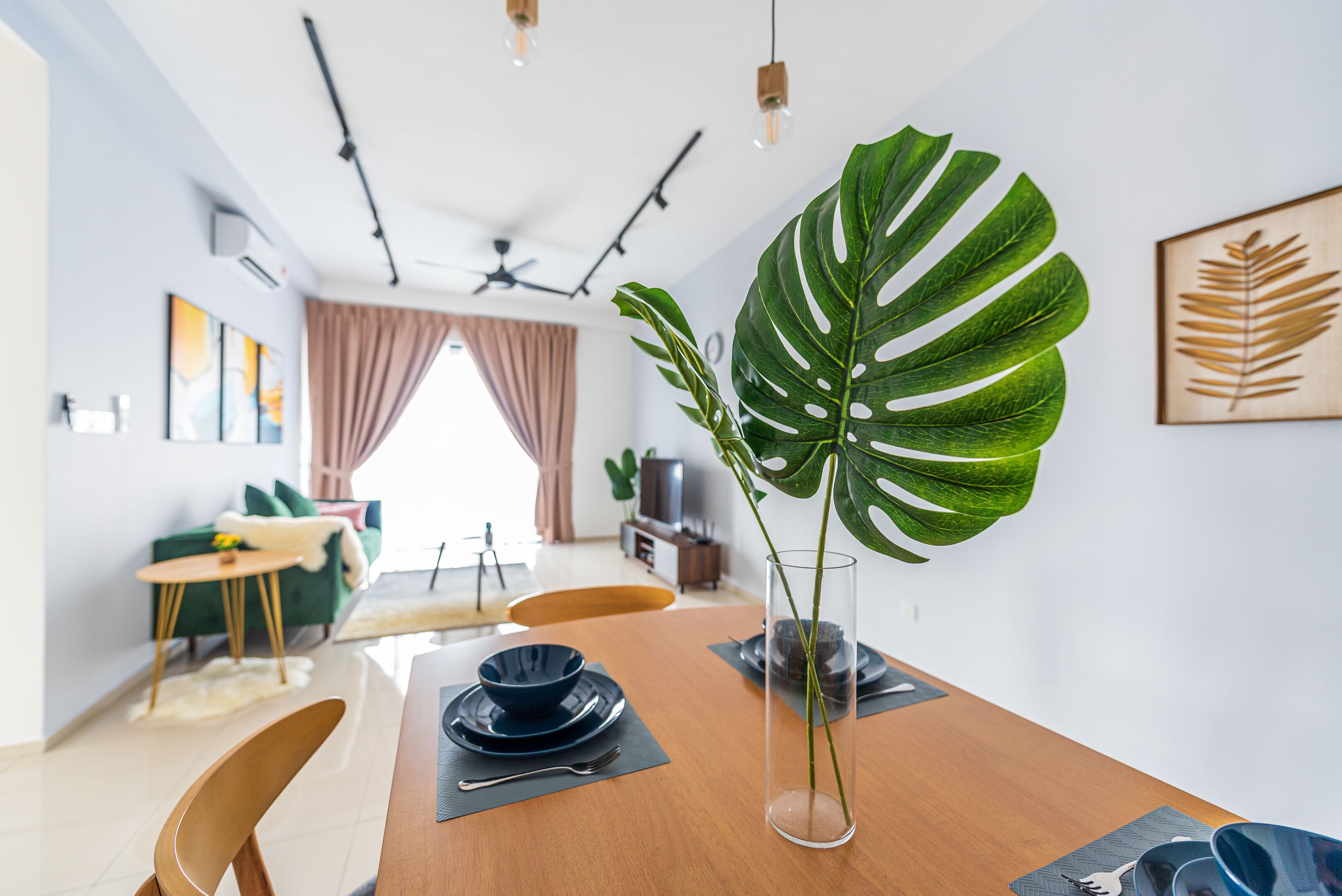 A dining area with a wooden table set for two, a large green plant, and a framed leaf artwork on the wall.