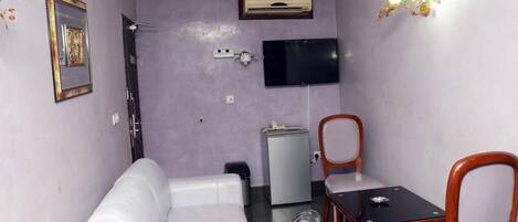 A small room with a white sofa, a table with two chairs, a television mounted on the wall, an air conditioning unit, and a small refrigerator.