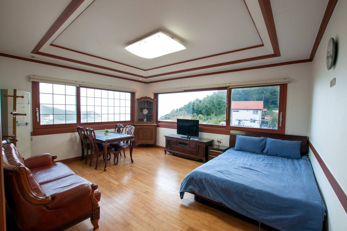 A bedroom with a bed, a TV, a dining table with chairs, and large windows offering a view of the outdoors.