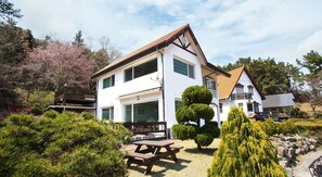Een wit gebouw met twee verdiepingen, een balkon, een terras met een tafel en stoelen, en een verzorgde tuin met verschillende struiken en bomen.