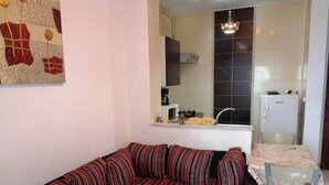 A living room with a striped sofa, a dining table with a chair, a kitchen area with a refrigerator, and a decorative wall piece. - Furnished Short Stay Apartment In Tunis (Tunis)