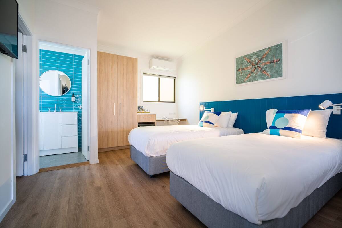 A hotel room with two beds, a wooden wardrobe, a bathroom with blue tiles, and a painting on the wall.