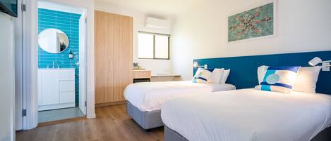 A hotel room with two beds, a wooden wardrobe, a bathroom with blue tiles, and a painting on the wall.