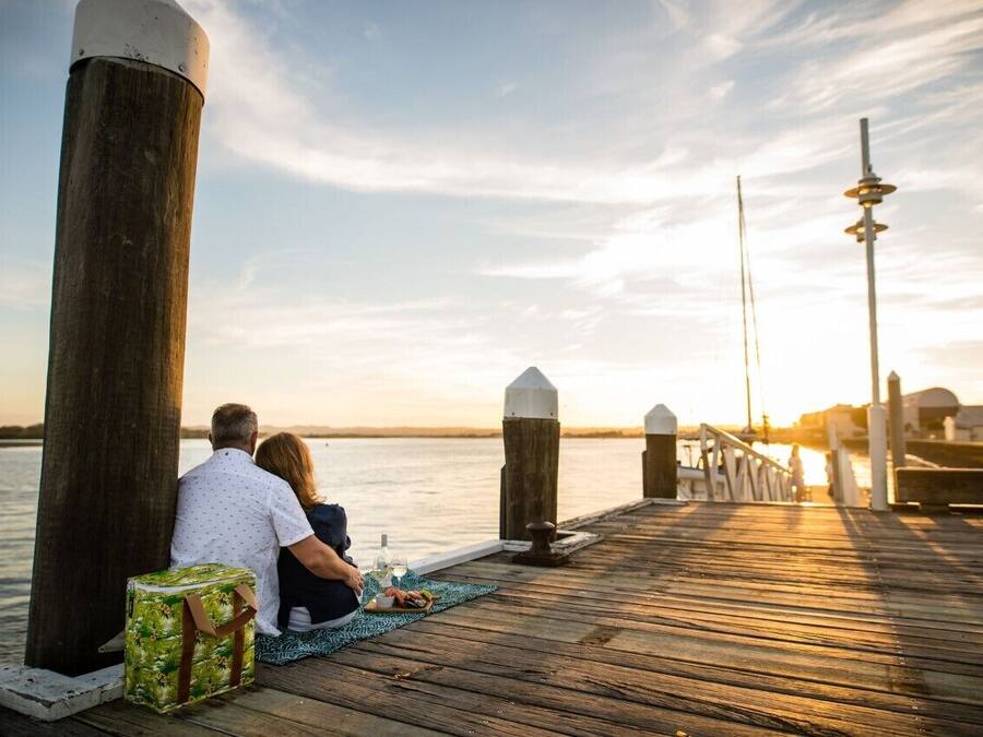 Una pareja sentada sobre un muelle de madera, con vista al agua y un atardecer de fondo.