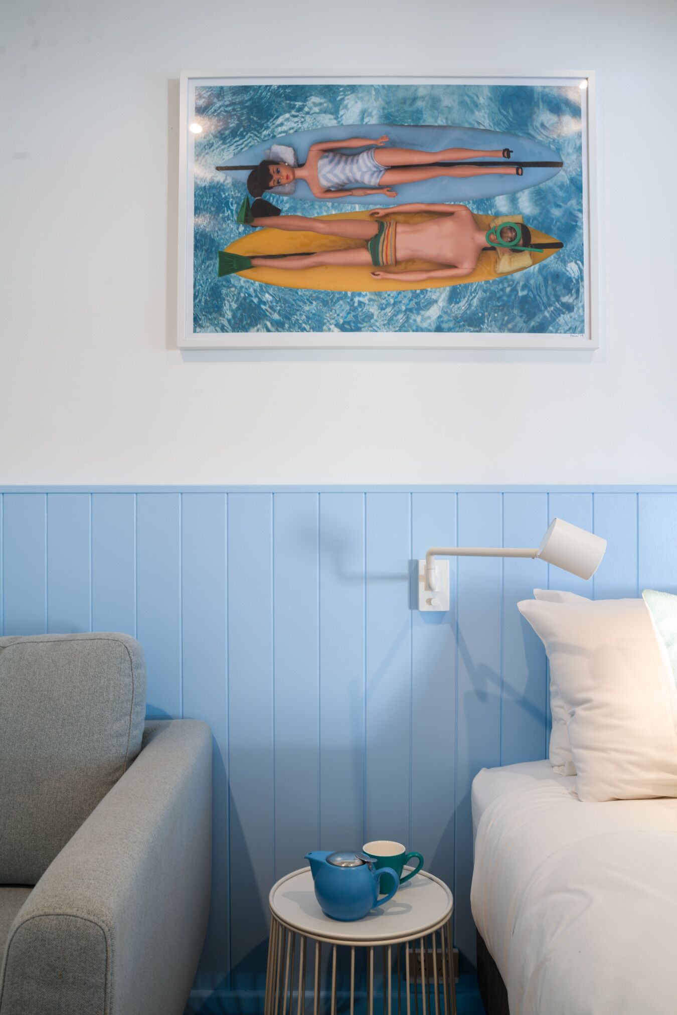 A room with a bed, a small round table with a blue mug, a grey sofa, and a painting of two people in a boat on a blue background.