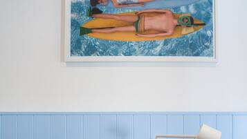 Une pièce comprenant un lit, une petite table ronde avec une tasse bleue, un canapé gris et un tableau représentant deux personnes dans un bateau sur un fond bleu.