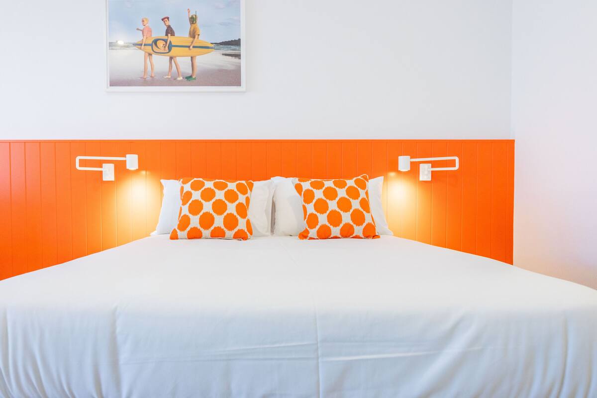 A bed with white bedding and orange accents, a framed picture of people with surfboards on the wall, and a bright orange headboard.