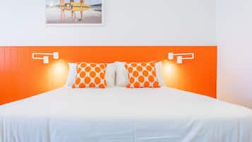 A bed with white bedding and orange accents, a framed picture of people with surfboards on the wall, and a bright orange headboard.