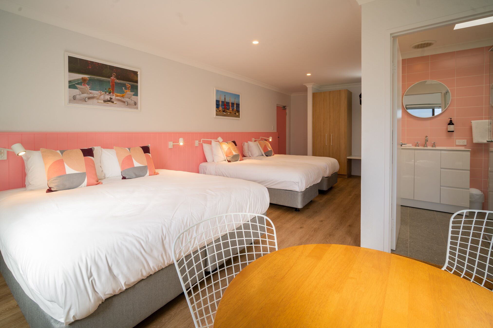 A hotel room with two beds, a wooden table, and a bathroom with pink tiles.