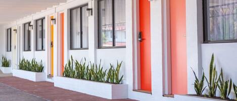 Una hilera de habitaciones de hotel con paredes blancas y puertas naranjas, cada una con ventana y una pequeña maceta con plantas.
