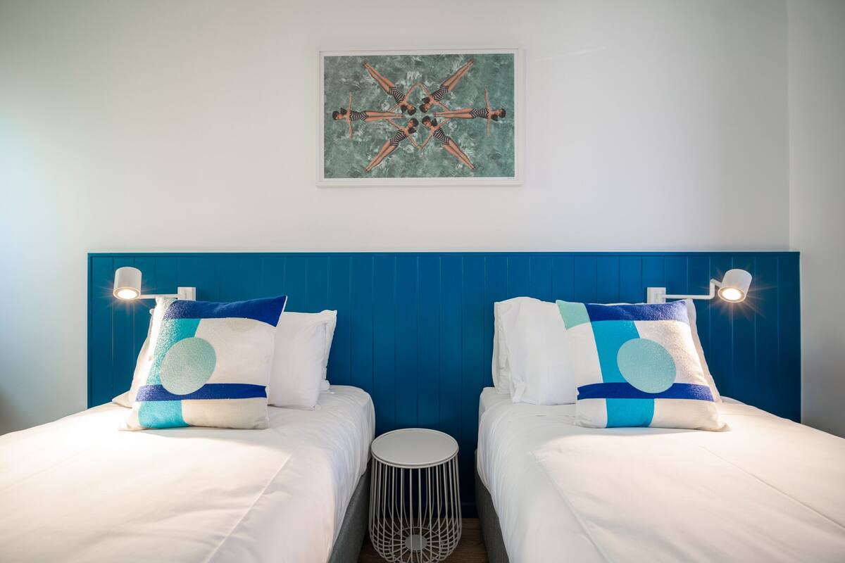 A hotel room with two beds, a blue headboard, a small round table, and a wall-mounted artwork.