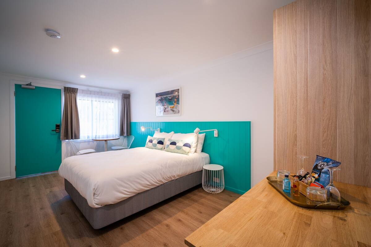 A modern hotel room with a teal accent wall, a bed with white bedding, a small round table with a tray of amenities, and a window with curtains.