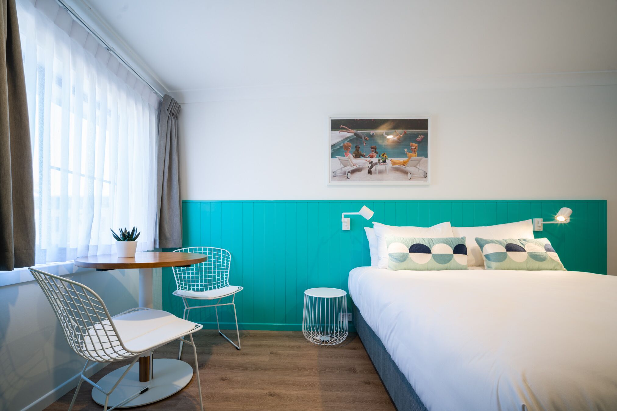 Une chambre d’hôtel moderne avec un mur d’accent de couleur turquoise, un grand lit agrémenté d’oreillers à motifs, une petite table ronde avec des chaises, une plante en pot et un tableau encadré au mur.