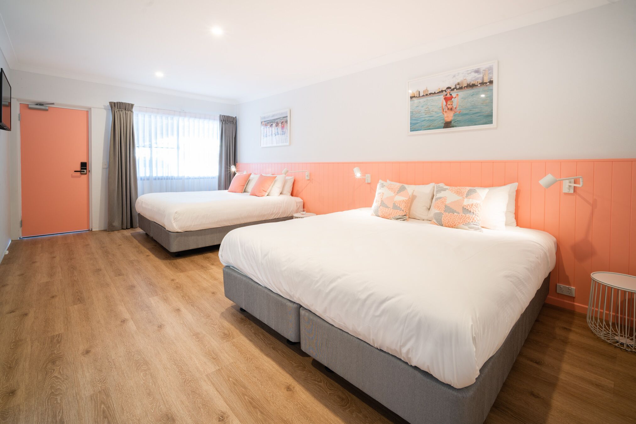 A hotel room with two beds, wooden flooring, a door, and a window with curtains.