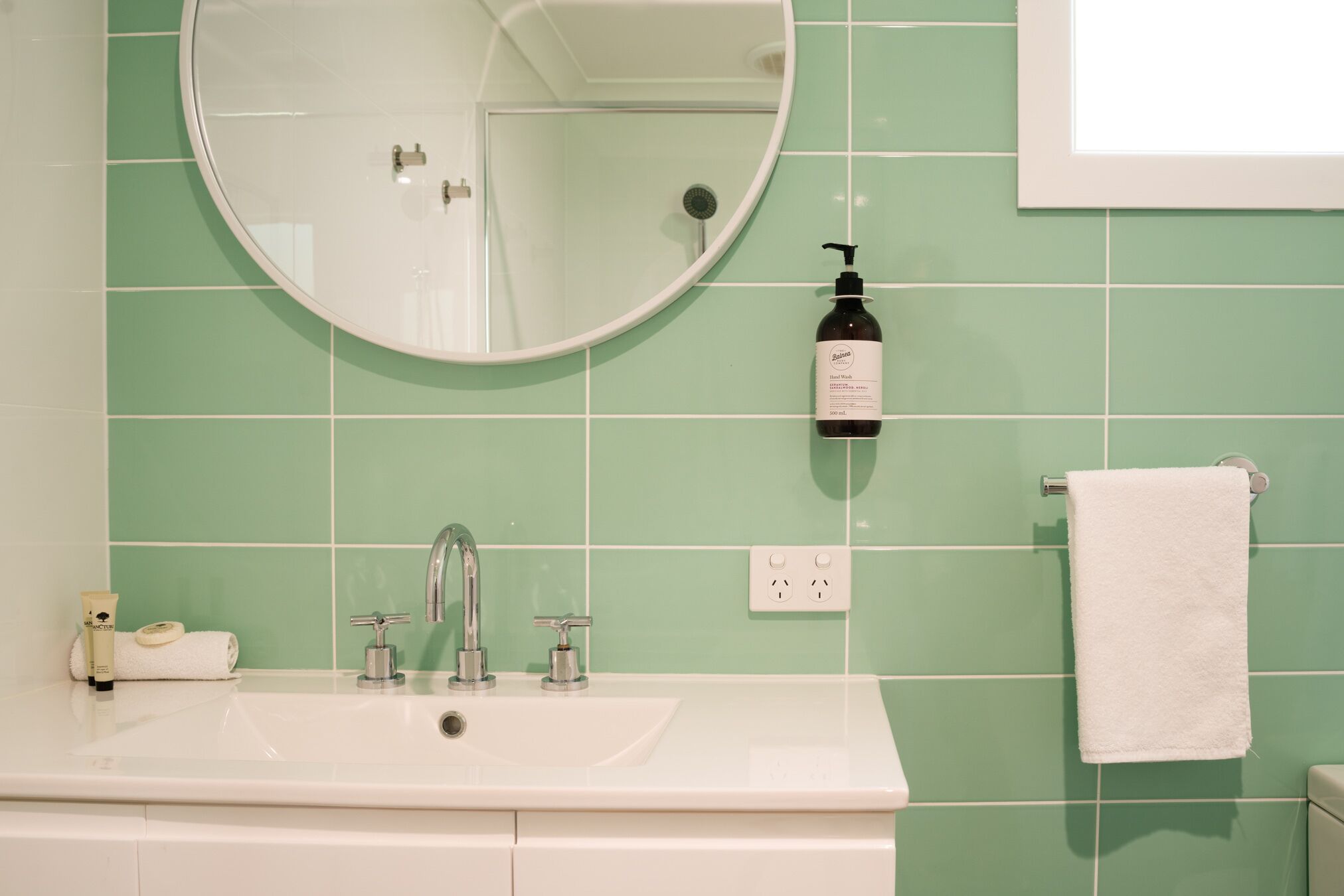 A bathroom with a white sink, a round mirror, a handheld showerhead, a bottle of shampoo, and a white towel hanging on a rack.