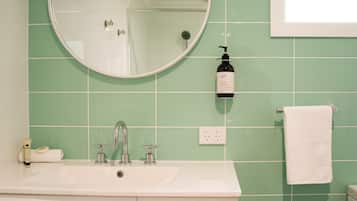 Un bagno con un lavandino bianco, uno specchio rotondo, un soffione doccia manuale, una bottiglia di shampoo e un asciugamano bianco appeso a un porta-asciugamani.