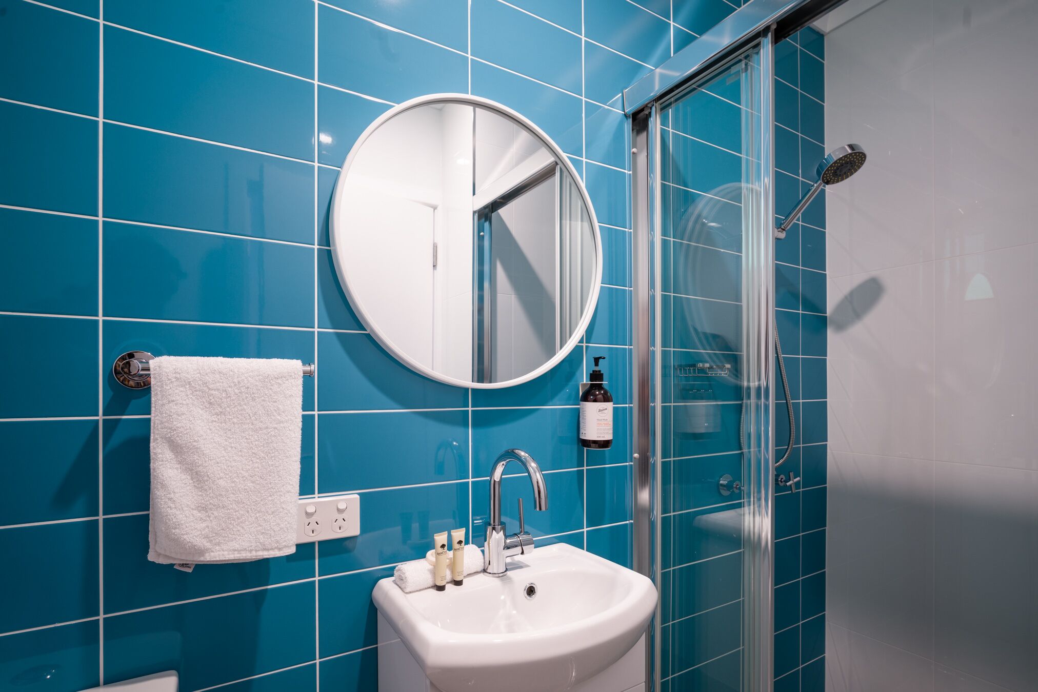 Une salle de bain avec des carreaux bleus, un miroir rond, un lavabo blanc, une douche en verre et une serviette blanche accrochée au mur.