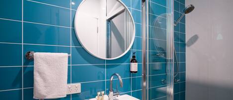 Une salle de bain avec des carreaux bleus, un miroir rond, un lavabo blanc, une douche en verre et une serviette blanche accrochée au mur.