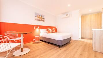 A hotel room with a bed, a dining table, and a chair. There is a picture on the wall and an air conditioning unit.