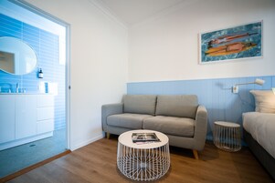 Un dormitorio moderno con un sofá gris, una mesita redonda, una cama con ropa de cama blanca, una mesita de noche pequeña, una lámpara de pared y un cuadro de un barco en la pared.