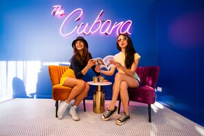 Two people sitting on a couch in a room with a neon sign reading "The Cubana".
