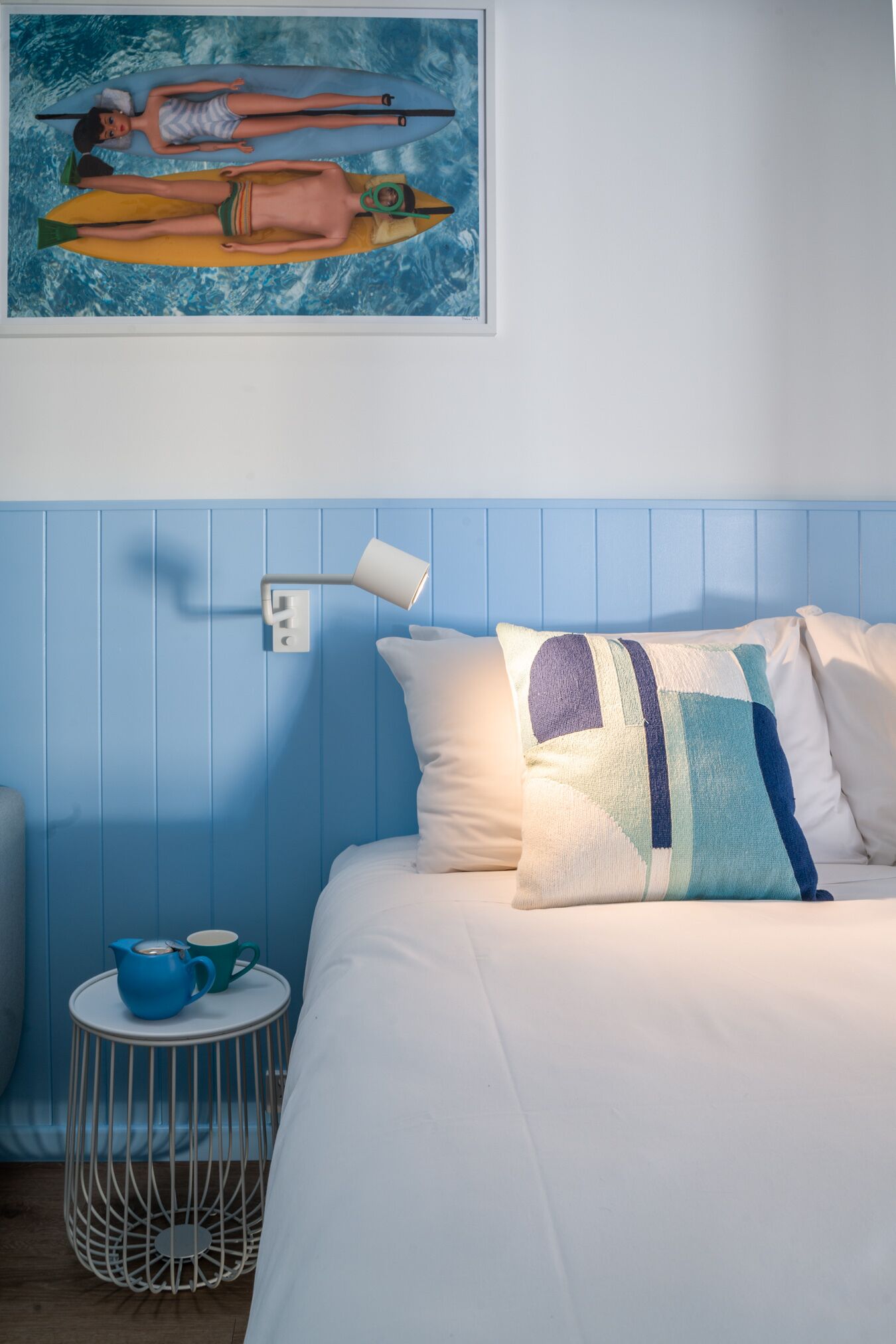 Un dormitorio con cama, almohadas, una lámpara de pared, un cuadro y una pequeña mesa con tetera y taza.