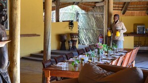 A dining area with a wooden table set for four, surrounded by chairs. There is a statue on a shelf, a painting on the wall, and a person standing near a table with a yellow bottle. - Jumbo Junction (Gunotsoga)