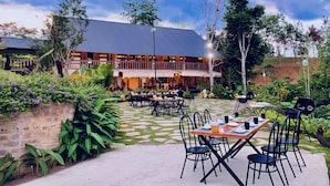An outdoor dining area with tables and chairs, surrounded by lush greenery and a building with a balcony.