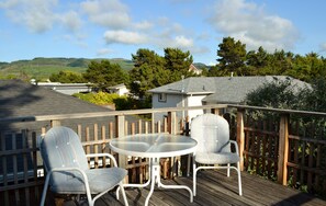 Une terrasse en bois avec deux chaises et une table, entourée d’une clôture en bois, donnant sur un quartier résidentiel avec des maisons et des collines.