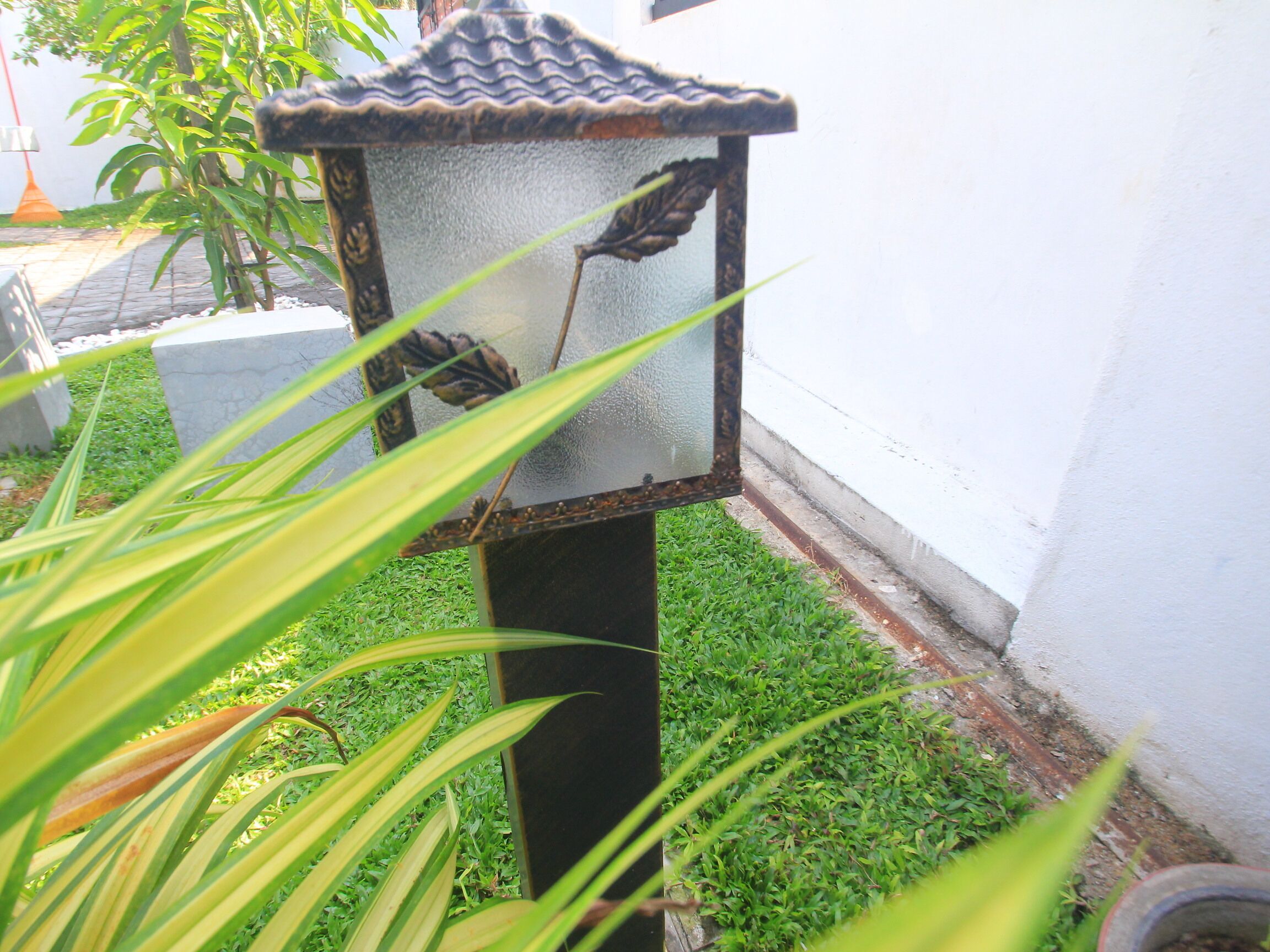 Una linterna tradicional con un diseño decorativo, ubicada sobre una base de concreto en un jardín con césped y una pared blanca al fondo.